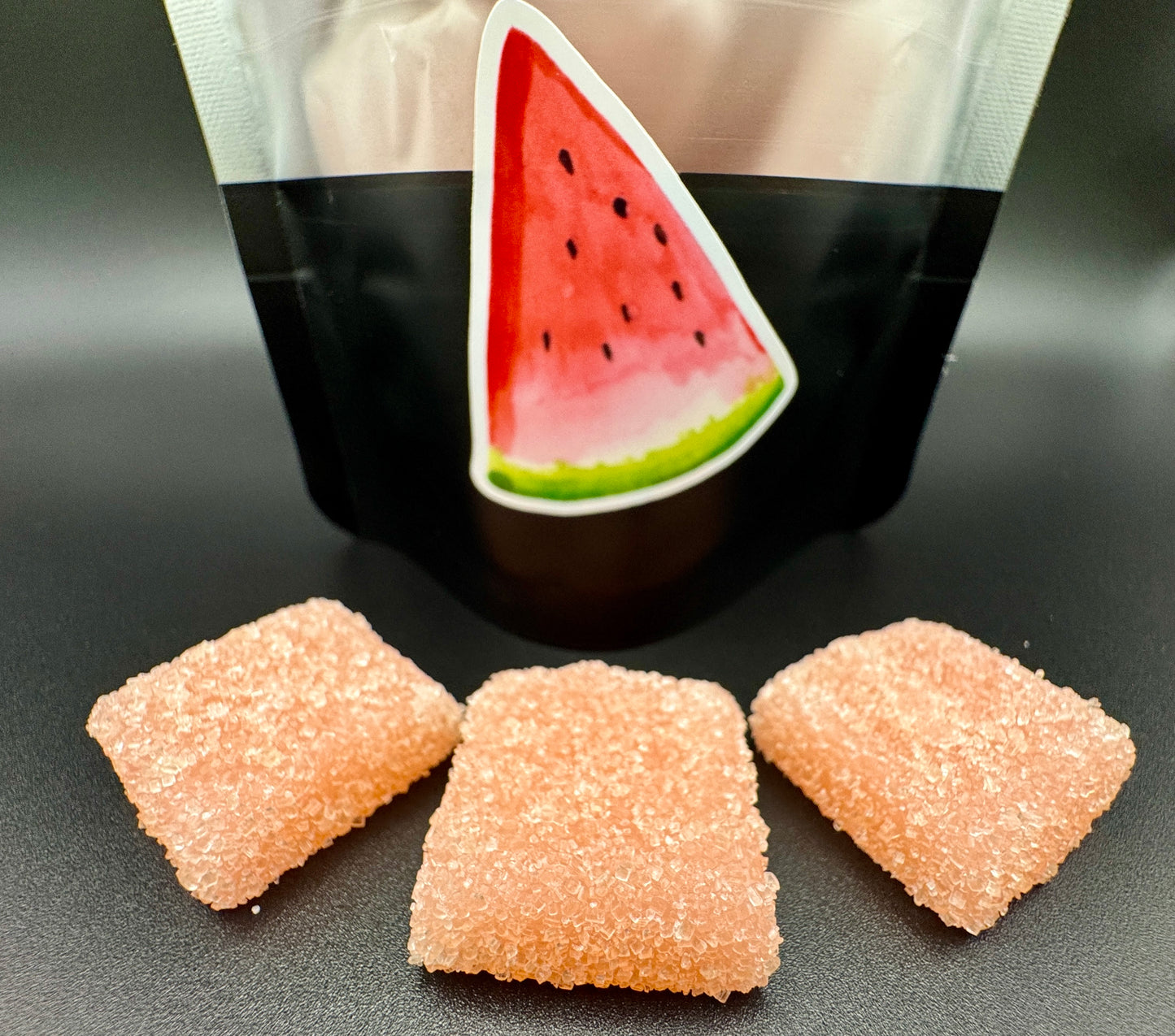 Three pink gummy candies in front of a black and white bag with a watermelon design.