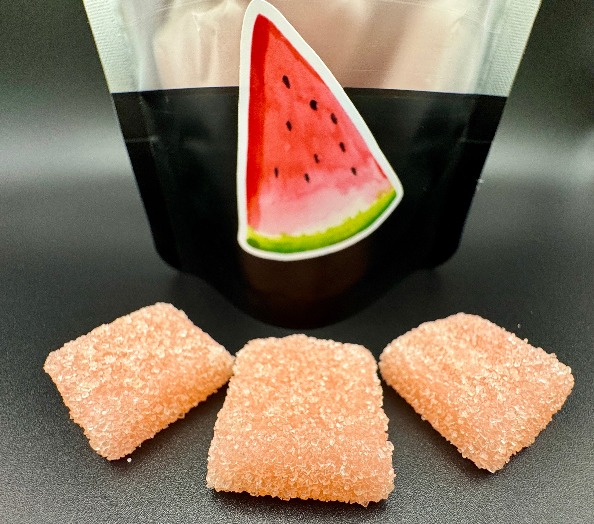 Three pink gummy candies in front of a black and white bag with a watermelon design.